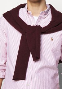 Burgundy stickad tröja knuten över en rosa och vit randig skjorta med knappar; synlig logotyp på skjortan och mjuk textur på tröjan.