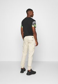 T-shirt noir avec des manches vertes et blanches à motifs, associé à un pantalon cargo beige. Bottes noires avec une finition texturée.