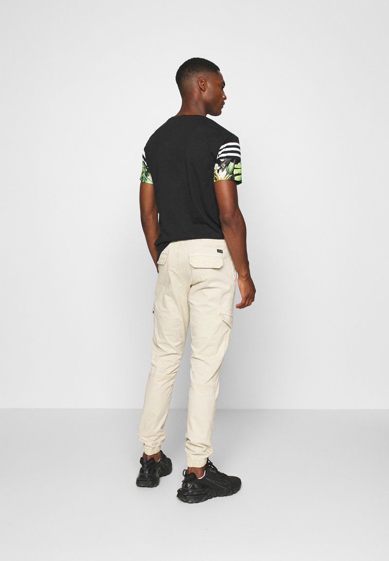Camiseta negra con mangas a rayas en verde y blanco, combinada con pantalones cargo beiges. Las botas son negras con un acabado texturizado.