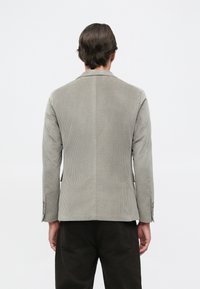 Šedý kordurový blazer se strukturovaným designem ramen, jedním roz ventilačním otvorem a knoflíkovými manžetami; nošený přes tmavé kalhoty. Zadní pohled.