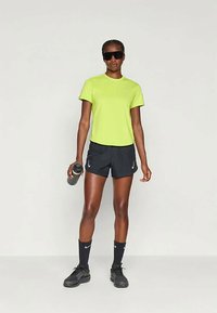 Neonsko zelena športna majica, črne kratke hlače, črne športne copate in črne nogavice do kolen. Drži sivo plastenko, nosi temna sončna očala.