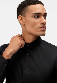Camicia nera a maniche lunghe, realizzata in tessuto liscio, con colletto a punta. Presenta bottoni neri e una silhouette aderente.