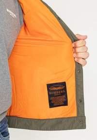 Zielona kurtka bombowiec z pomarańczowym podbiciem, z czarną metką z białym napisem. Gładka faktura i zapięcie na napy.