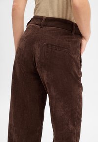 Persoon in een hoog- taille donkere bruine corduroy broek en een beige geribbelde top, met één hand bij de heup.