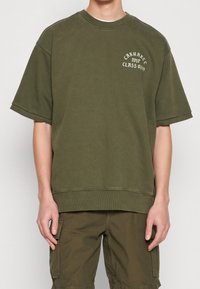 Sudadera oversized verde oliva, mezcla de algodón, cuello redondo acanalado, mangas cortas, con logotipo de texto blanco en el lado izquierdo del pecho.