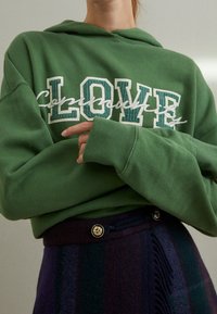 Groene hoodie met witte en teal letters die "LOVE" zeggen en het woord "community" in een cursieve lettertype. Zachte stof.