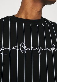 Černý svetr s bílými pruhy a vyšívaným kurzivním logem. Vybaven klasickým kulatým výstřihem a hladkou texturou.