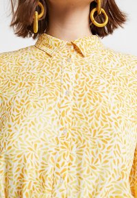 Žlutá a bílá košile na knoflíky s motivem listů. Měkká tkanina s límečkem a perlovými knoflíky, doplněná velkými kulatými náušnicemi.