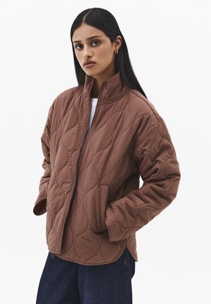 Ung kvinde med langt mørkt hår iført en brun quiltet jakke med lynlås og mørkeblå bukser, stående med hænderne i lommerne på jakken.