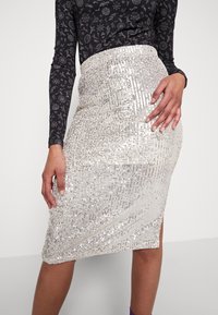 Silvrig paljettpencilkjol med en strukturerad yta. Har en åtsittande midja och är knälång, matchad med en svart mönstrad topp.