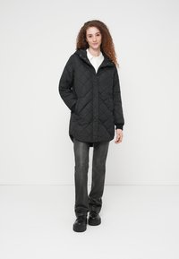 Μαύρο καπιτονέ μπουφάν με κουκούλα, με φερμουάρ και κούμπωμα με κουμπιά, πλαϊνές τσέπες και ελαφρώς επιμηκυμένη πλάτη. Φοριέται πάνω από λευκό πουκάμισο.