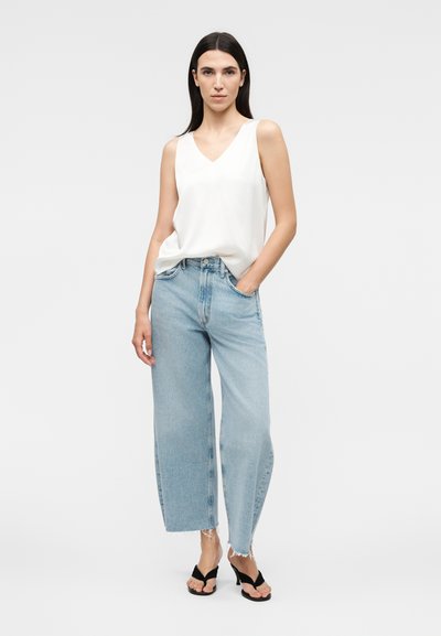 Γυναίκα στέκεται με το ένα χέρι στην τσέπη, φορώντας λευκό αμάνικο τοπ, ανοιχτό μπλε τζιν με φαρδιά μπατζάκια και μαύρα σανδάλια με τακούνι.