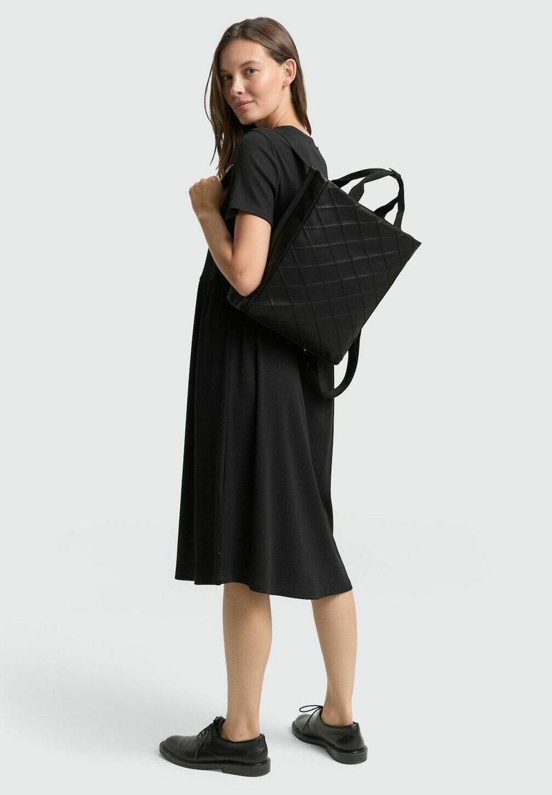 Schwarze, gesteppte Tragetasche mit doppelt getragenen Schulterriemen, die ein geometrisches Muster zeigt. Trägt man über einem schwarzen Kleid, das ein minimalistisches Design zur Schau stellt.