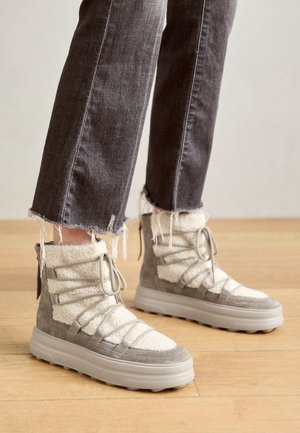 Voeten met grijze en witte winterlaarzen met veters en een fleece voering, gecombineerd met donkergrijze jeans met rafelige zoom, op een lichte houten vloer.