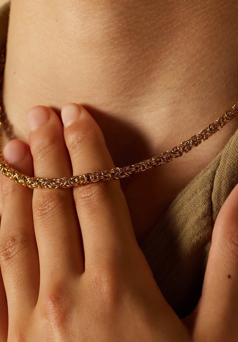 Gouden ketting met ingewikkelde schakels, rustend op de huid. Soepele textuur, close-up weergave benadrukt het ontwerp en de details van de sieraden.