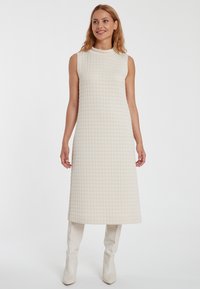 Ärmelloses cremefarbenes Kleid mit einem strukturierten Rautenmuster, knielang und mit hohem Kragen, kombiniert mit kniehohen weißen Stiefeln.