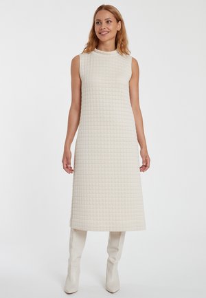 Robe sans manches crème avec un motif en grille texturé, à longueur genou et col haut, associée à des bottes blanches jusqu'au genou.