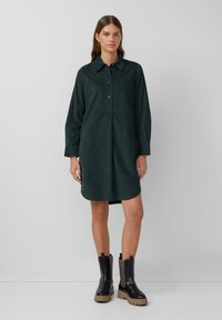 Abito camicia button-up verde scuro con maniche lunghe, tasche laterali e taglio dritto. Indossato con stivali neri chunky con accenti testurizzati.