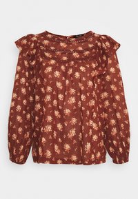 Blouse florale marron avec des épaules à volants, des manches longues et un tissu texturé. Présente de petits motifs de fleurs crème et rose sur un fond sombre.