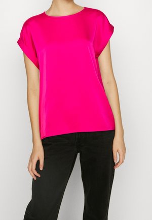 Mujer con blusa de manga corta de color rosa brillante combinada con pantalones negros, de pie frente a un fondo blanco liso.