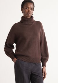 Maglione a collo alto oversized marrone con polsini e orlo a coste, in tessuto a maglia testurizzato, abbinato a pantaloni a righe.