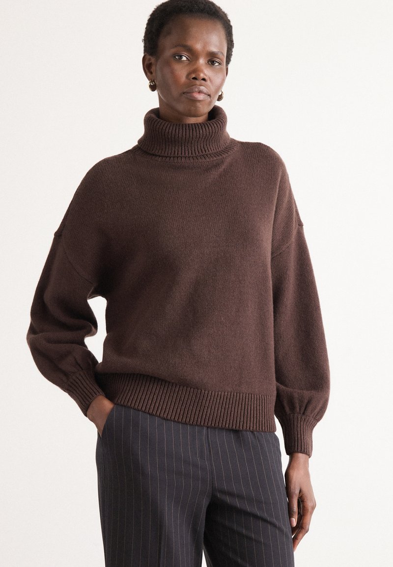 Maglione a collo alto oversized marrone con polsini e orlo a coste, in tessuto a maglia testurizzato, abbinato a pantaloni a righe.