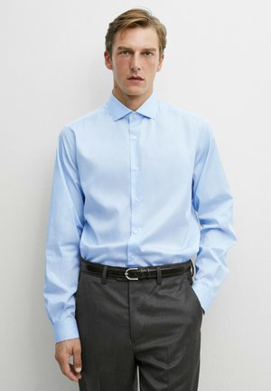 Uomo che indossa una camicia azzurra con bottoni infilata in pantaloni grigio scuro con cintura nera, in piedi davanti a uno sfondo bianco uniforme.