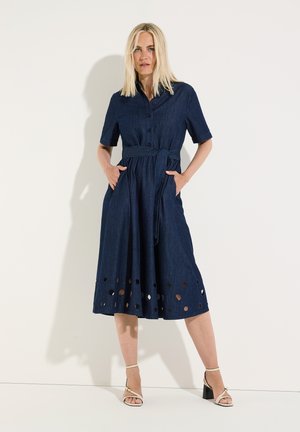 Woman standing against a white wall wearing a navy belted dress with short sleeves, cut-out hem, and white heeled sandals, hands in pockets.