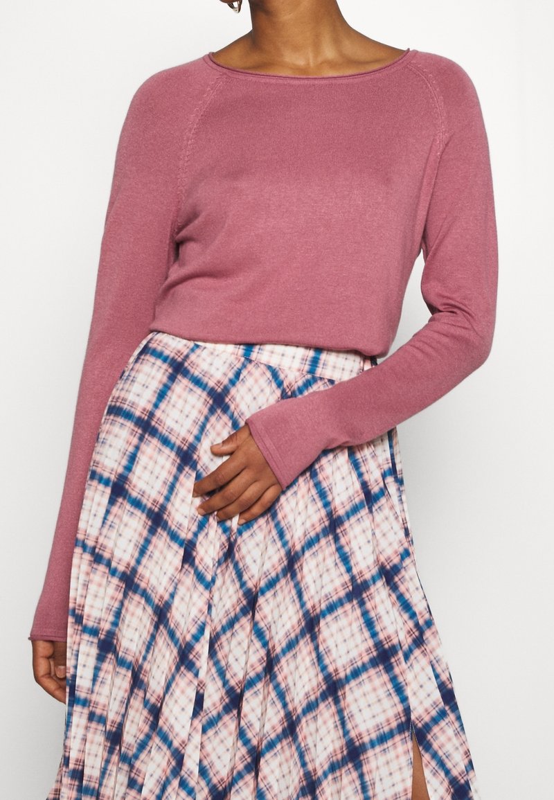 Femme portant un pull à manches longues rose poudré et une jupe taille haute à carreaux dans des tons bleus et roses avec une fente à l'avant.
