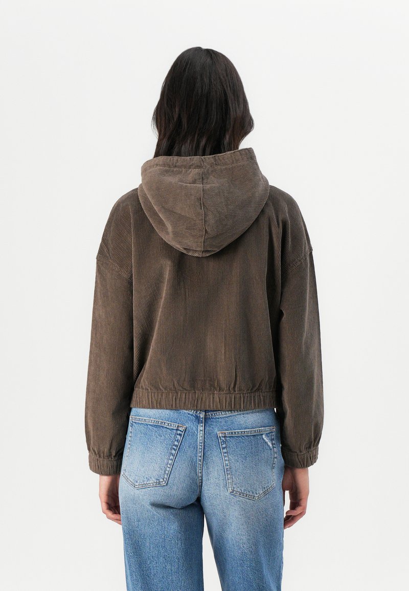 Veste cropped en velours côtelé marron avec une capuche, taille élastique et coupe décontractée. Associée à un jean bleu délavé, mettant en avant le design du dos.