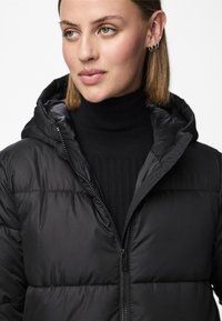 Juoda pūkinė striukė su užtrauktuku ir gaubtu. Turi juostas primenantį pasiuvimą ir lygų, blizgantį paviršių. Dėvima ant juodo riboto aukštu kaklu megztinio.