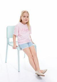 Camiseta de algodón rosa con "esperanza" en cursiva, combinada con pantalones cortos de mezclilla. Chica sentada en una silla azul pastel, usando zapatos de color beige claro.
