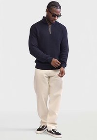 Mies, joka pitää yllä mustaa ribattua neuletta vetskarilla, vaaleita housuja, mustia ja valkoisia tennareita sekä aurinkolaseja, säätää neuleensa hihaa.