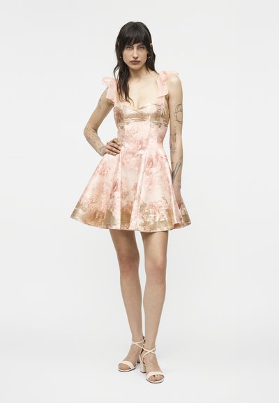 Femeie cu păr închis la culoare, purtând o rochie florală roz și aurie cu bretele cu volane și sandale bej cu barete, stând cu o mână pe șold.