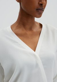 Chemisier blanc avec un décolleté en V, fabriqué à partir d'un tissu texturé léger. Présente un design épuré et une drape douce.