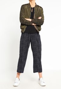 Osoba stojąca z założonymi ramionami, ubrana w oliwkową bomberkę, czarną bluzkę, wzorzyste szerokie spodnie oraz białe sneakersy.