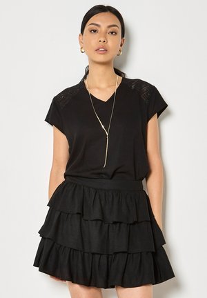 Kobieta w czarnej bluzce z krótkim rękawem z koronkowymi detalami na ramionach oraz czarnej spódnicy z falbanami ułożonymi warstwowo, ozdobiona złotymi kolczykami kołami i długim naszyjnikiem.