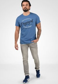 Modra bombažna majica s črnim napisom "Pravilno originalne mešanice denim blaga", v paru s svetlo sivo obleko in modrimi supergami.