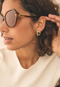 Solglasögon med sköldpaddsmönster och en djärv, kantig form med bruna linser. Guldiga örhängen med en vriden design. Texturerad vit ribbad topp.