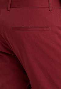 Pantalons bordeaux avec une texture lisse et une poche arrière unique, présentant des coutures discrètes. La taille est plate et ajustée.