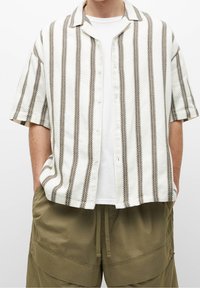 Camisa de manga corta blanca con rayas verticales marrones. Tejido texturizado con botones en la parte delantera y cuello clásico. Lleva una camiseta blanca debajo.