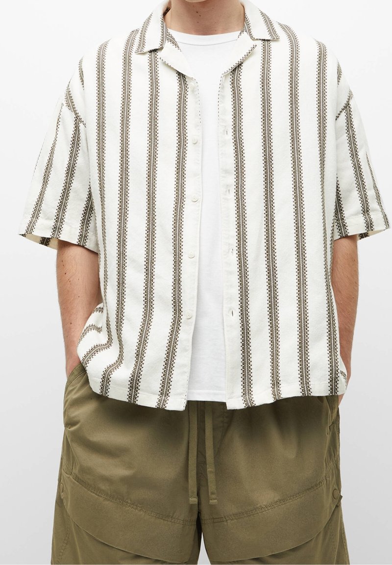 Koszula z krótkim rękawem w białym kolorze z brązowymi pionowymi paskami. Tkanina o fakturze, z przodem na guziki i klasycznym kołnierzykiem. Nosi się ją na białej koszulce.
