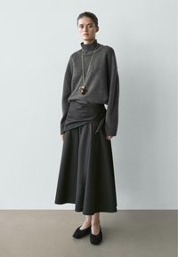 Grijze coltrui, zwarte wikkelrok, zwarte ballerina's. Zachte textuur, losse pasvorm, lange mouwen, en een gouden ketting met een hanger.