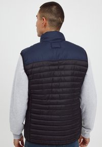 Marineblauer und schwarzer gepolsterter Weste mit hohem Kragen, die horizontale gesteppten Muster aufweist und einen Reißverschluss an der Front hat, getragen über einem grauen Pullover.