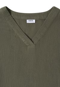 Haut en tissu texturé vert olive avec encolure en V et étiquette blanche indiquant "CECIL" au col.
