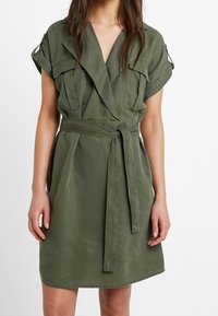 Abito avvolgente verde oliva realizzato in tessuto morbido con maniche corte, vita con cintura e tasche laterali discrete, caratterizzato da un design semplice.