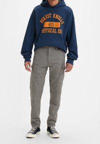 Marinblå hoodie med orange text, i kombination med grå cargobyxor med sidofickor och avsmalnande passform, tillsammans med svarta sneakers med vita sulor.