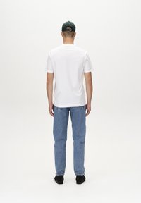 T-shirt bianca a maniche corte, vestibilità rilassata, abbinata a jeans in denim azzurri. Scarpe nere e cappellino verde completano l'outfit. Sfondo minimalista.