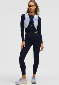 Leichte graue Hydrationsweste über einem enganliegenden schwarzen Langarmshirt und hochgeschnittenen schwarzen Leggings. Graue und orange Laufchuhe.