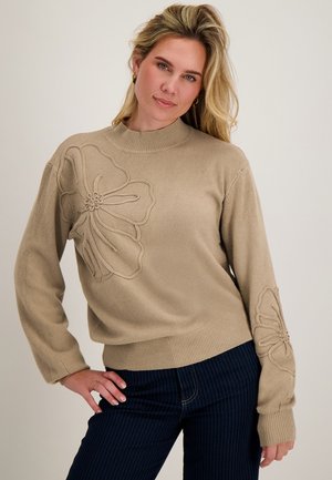 Vrouw met lang blond haar draagt een beige trui met grote bloemenborduursel en donkere broek met fijne strepen, poseert met hand op de heup.